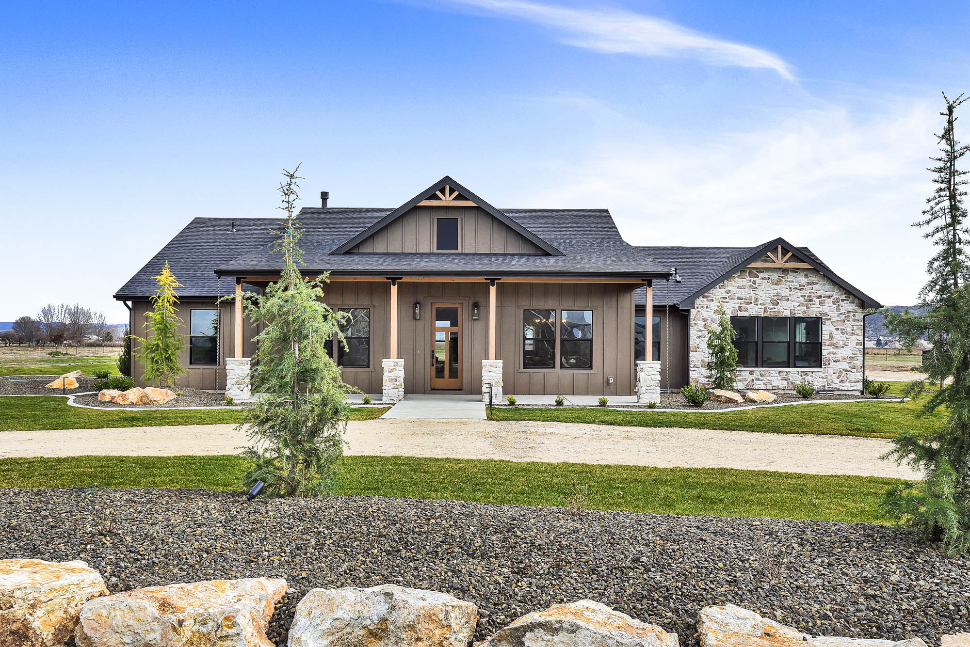 A brown house with a black roof and brick accents with a circle driveway set against a blue sky